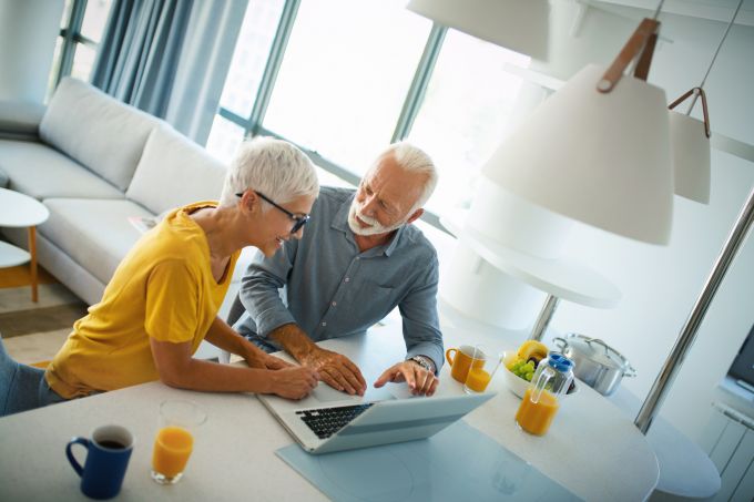 Two older adults smile and lean over a kitchen counter together, reviewing content on a laptop. A glass of orange juice and a coffee mug sit nearby in a bright, modern home.