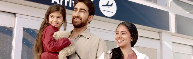 Illustration of a smiling newcomer family that is standing in front of a Canadian city skyline and holding a suitcase.