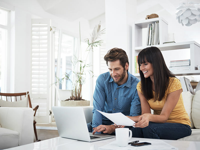 Couple sitting on their couch at home using a laptop while referring to a paper document