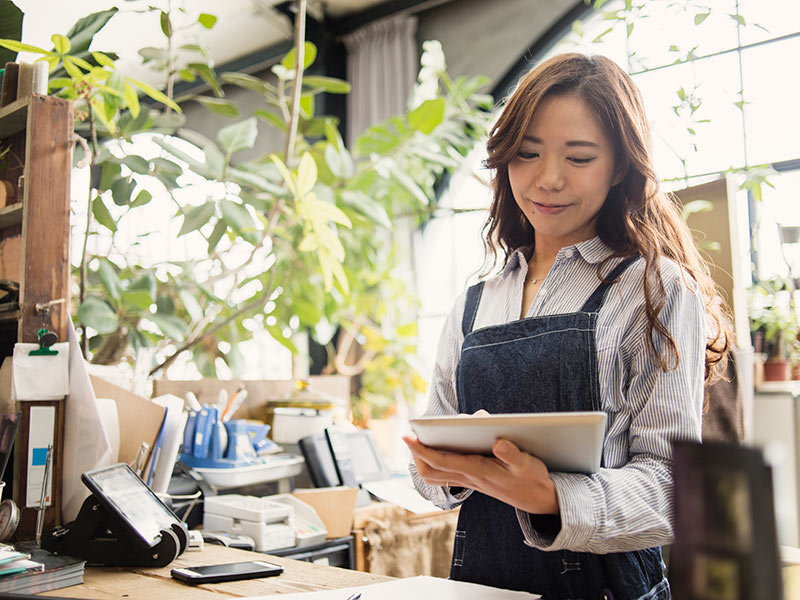 Woman wearing an apron standing behind a shop counter while using a tablet