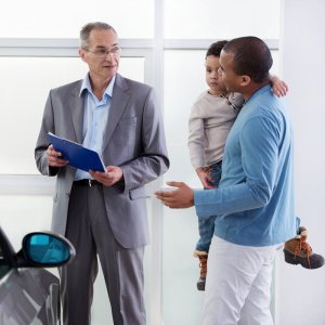 Car salesperson in a showroom showing a document to a man with a young child on his hip while standing next to a new car