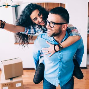 Woman playfully jumping on the back of a young man standing amongst boxes while she dangles the keys to their new home