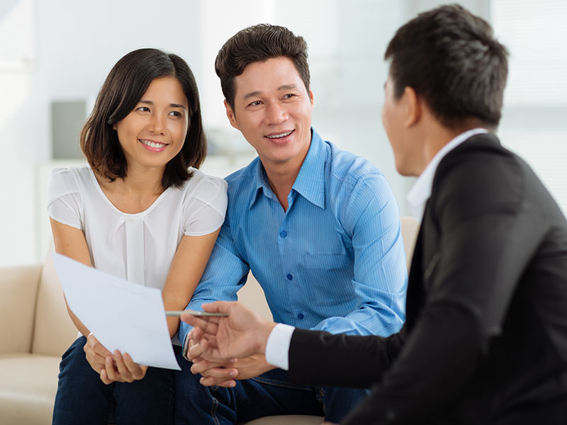 alt="Advisor pointing to a paper document with a pen while speaking to a couple sitting on a couch"