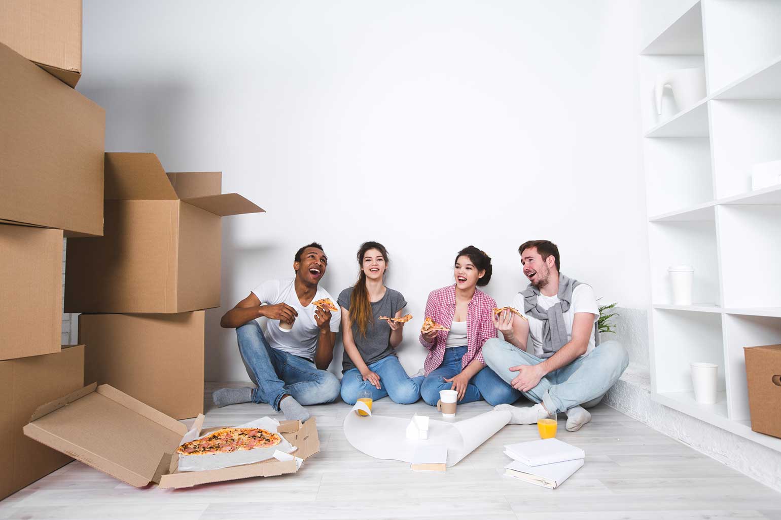 Happy first time home buyers and their 2 friends eating pizza on the floor after unpacking moving boxes.