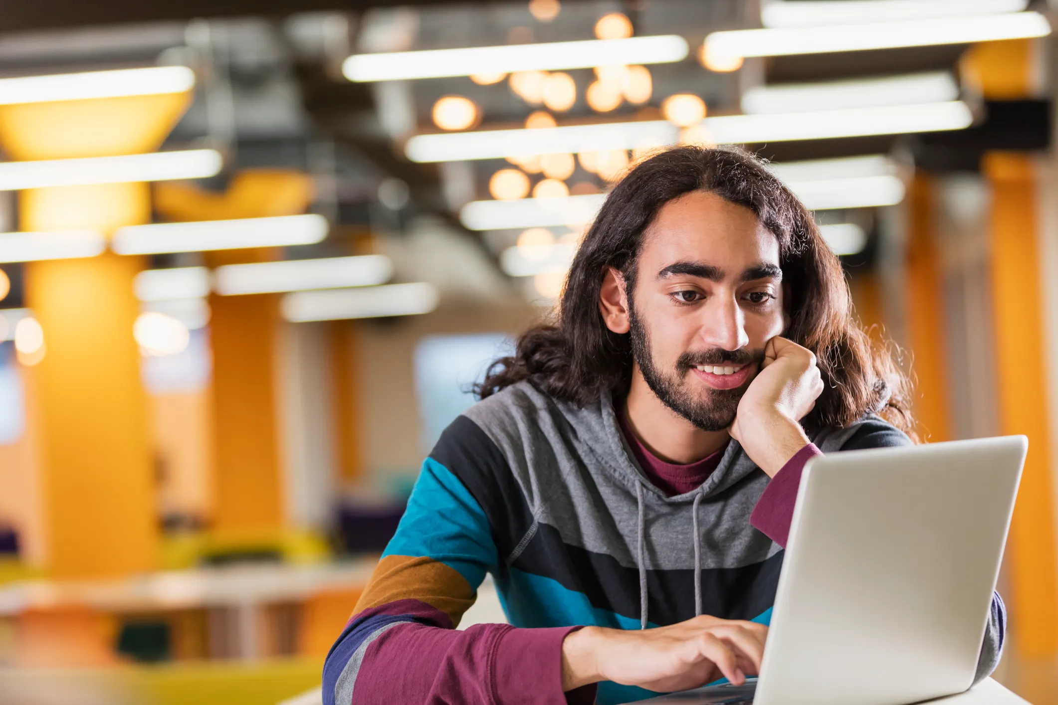 A young mixed race Indian and Caucasian man, in his 20s, with black hair, beard and mustache. He is working in an office wearing casual clothing, using a laptop computer.