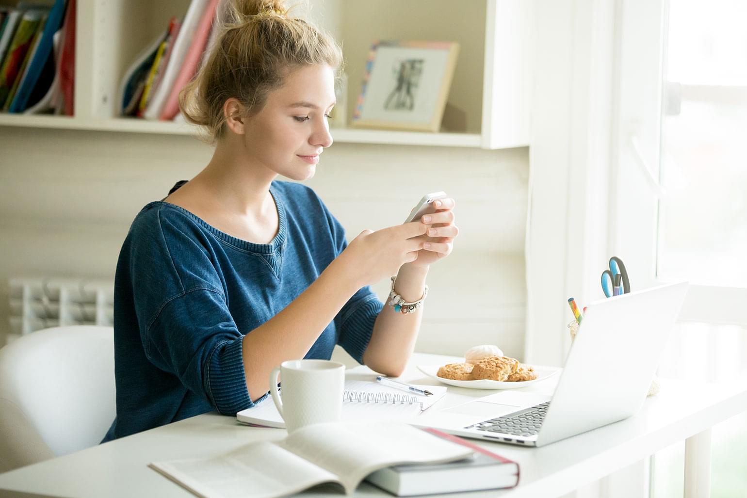 Portrait of young beautiful casual woman holding smartphone, looking at screen, using app or messaging while sitting at modern workplace with laptop, books, coffee and cookies in home office or dorm.
