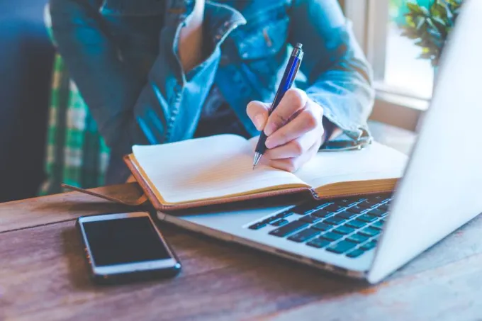 A person writing on a notebook in front of a laptop