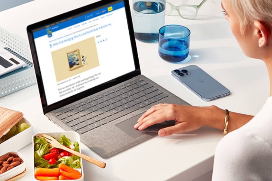 Woman sitting at a modern desk with a healthy lunch beside her in containers and her right hand using a laptop.