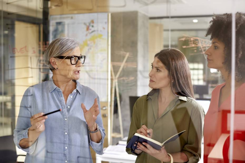 Three women are chatting near a glass whiteboard.