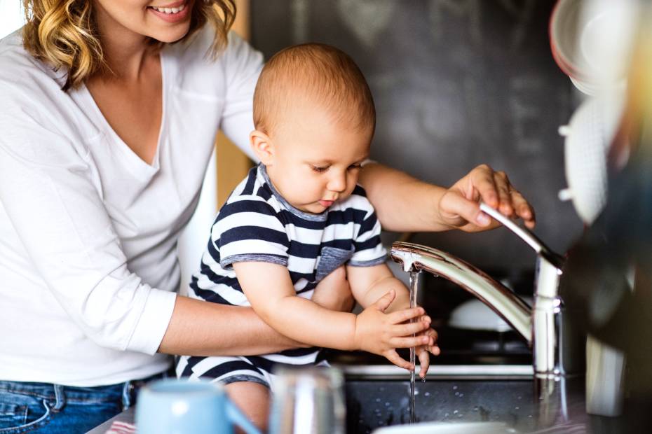 A parent holds their baby at the kitchen sink with the faucet running