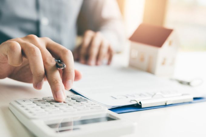 A person using a calculator and holding a pen at a desk, with a clipboard and a small model house in the background, representing the financial planning involved in buying a home.