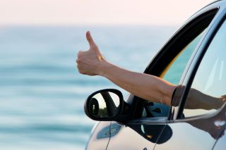 Driver in a car by the ocean displaying a thumbs up out his driver window
