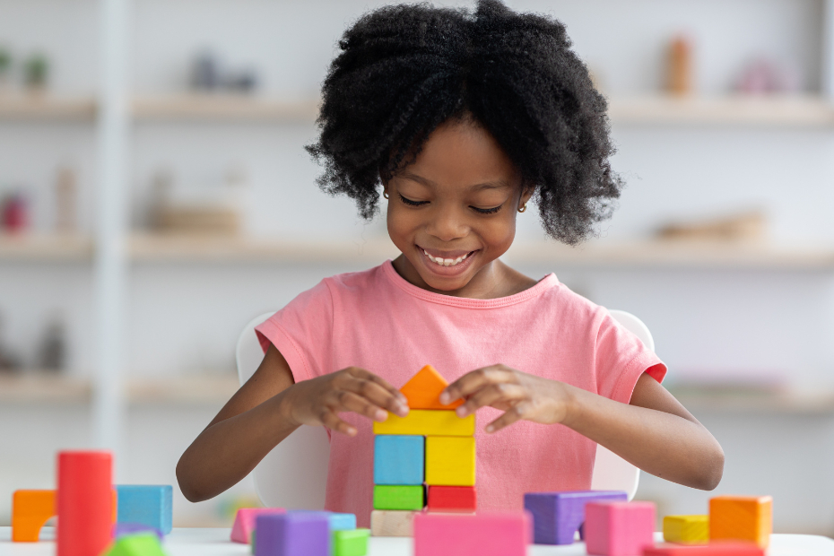 Happy young African American girl in bright pink shirt, playing with colorful wooden blocks