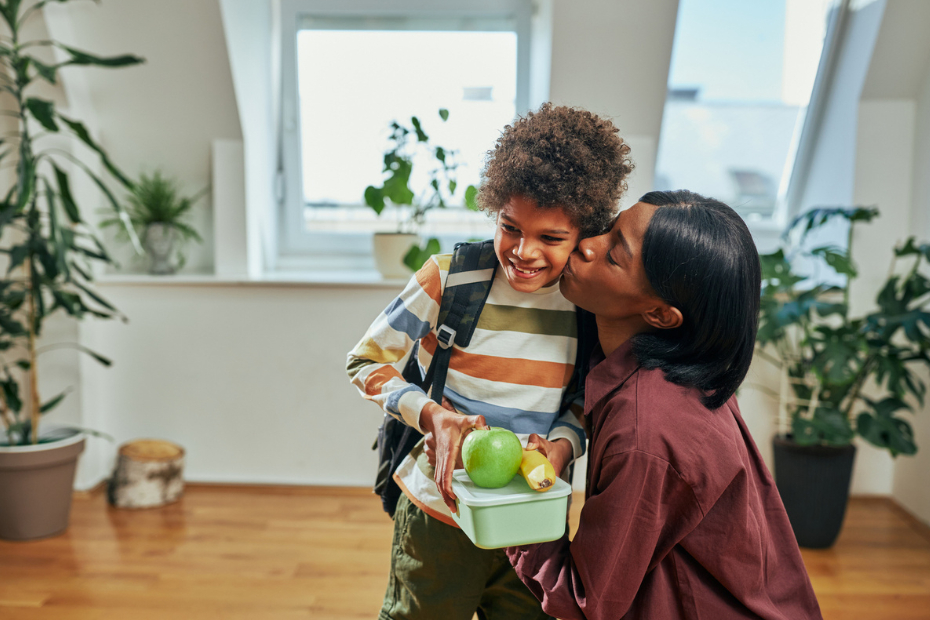 Woman hugging a young child holding plastic container, an apple and a banana.