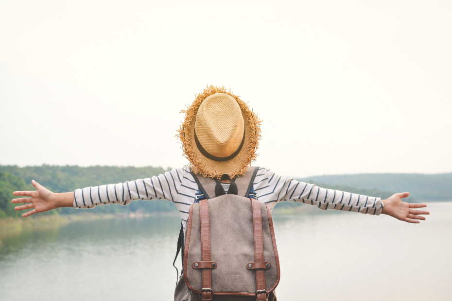 Young Asian girl backpacking by a lake with arms lifted up to the sky, in elation.