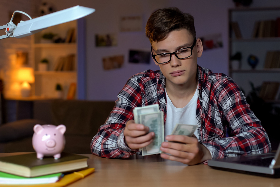 Teenage boy counting dollar bills at his desk, putting them to piggybank