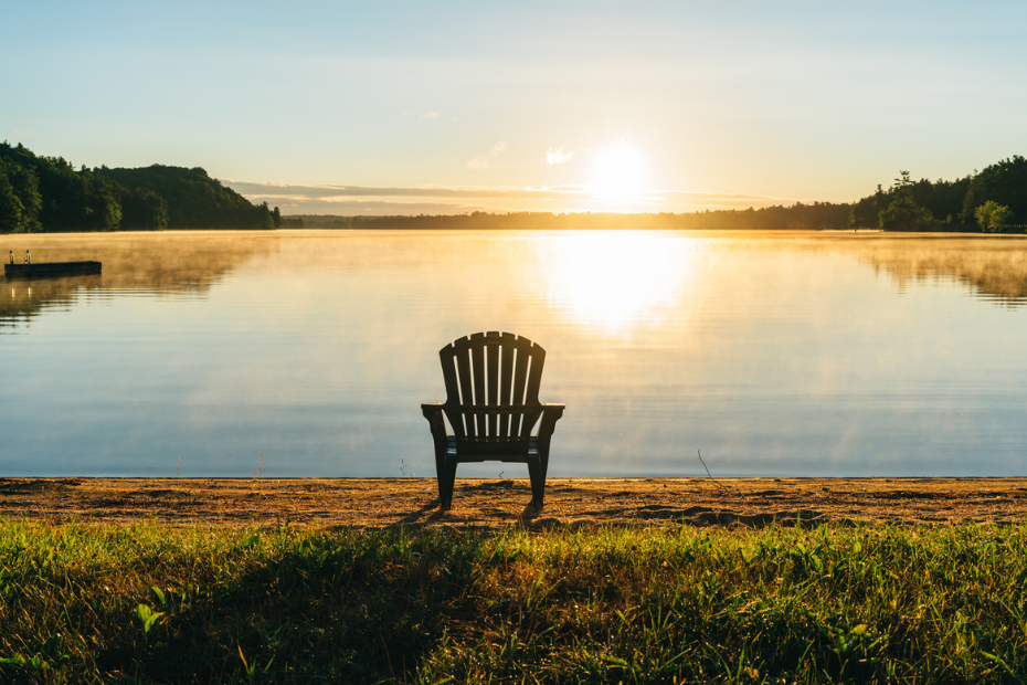 An empty Muskoka chair on a small beach beside a calm lake at sunset.