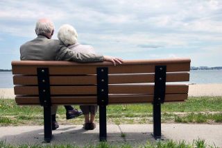 An elderly couple sitting together on a bench, looking out onto the waterfront.