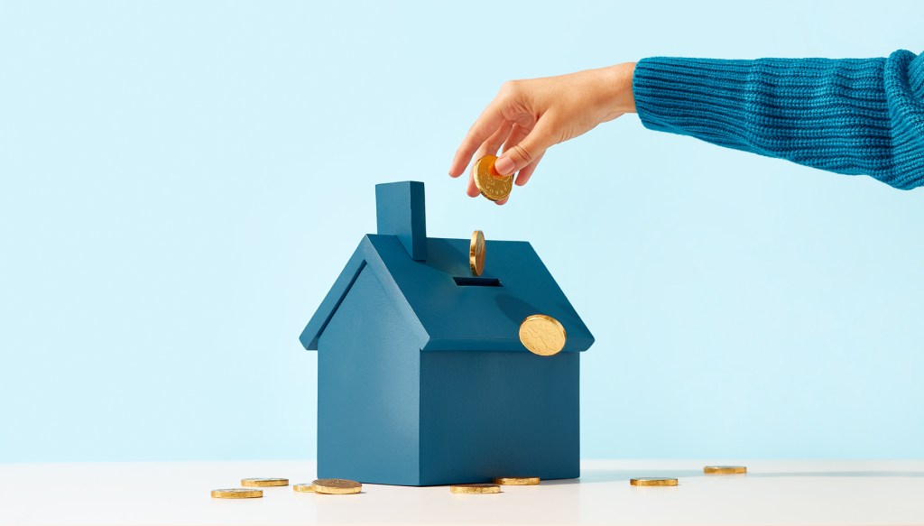 Hand dropping coins into a piggy bank shaped as a house.