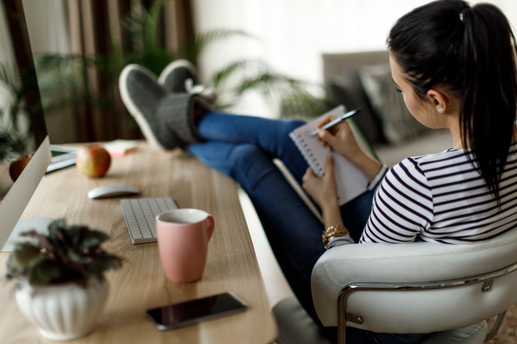 Woman facing away from the camera with her feet up on a desk and writing in a notebook.