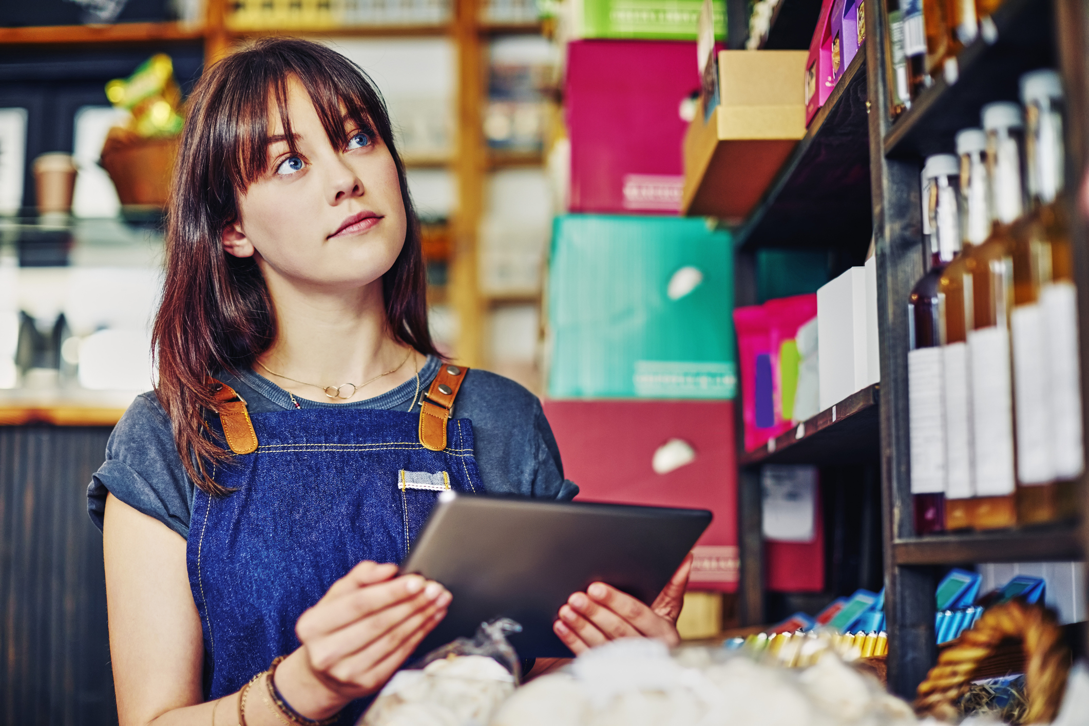 Beautiful saleswoman checking inventory with digital tablet in store. Confident deli owner is looking up while holding wireless computer in delicatessen. She is wearing apron.