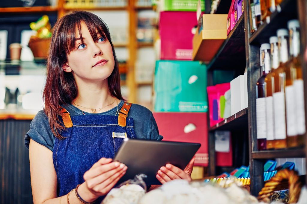 Beautiful saleswoman checking inventory with digital tablet in store. Confident deli owner is looking up while holding wireless computer in delicatessen. She is wearing apron.