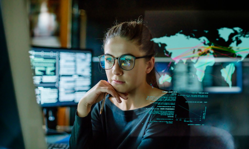 A young woman, wearing glasses, surrounded by computer monitors in a dark office. In front of her there is a see-through displaying showing a map of the world with some data.