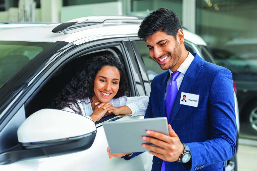 Lady sitting inside a new car at an auto dealership and having a conversation with a cheerful sales assistant.