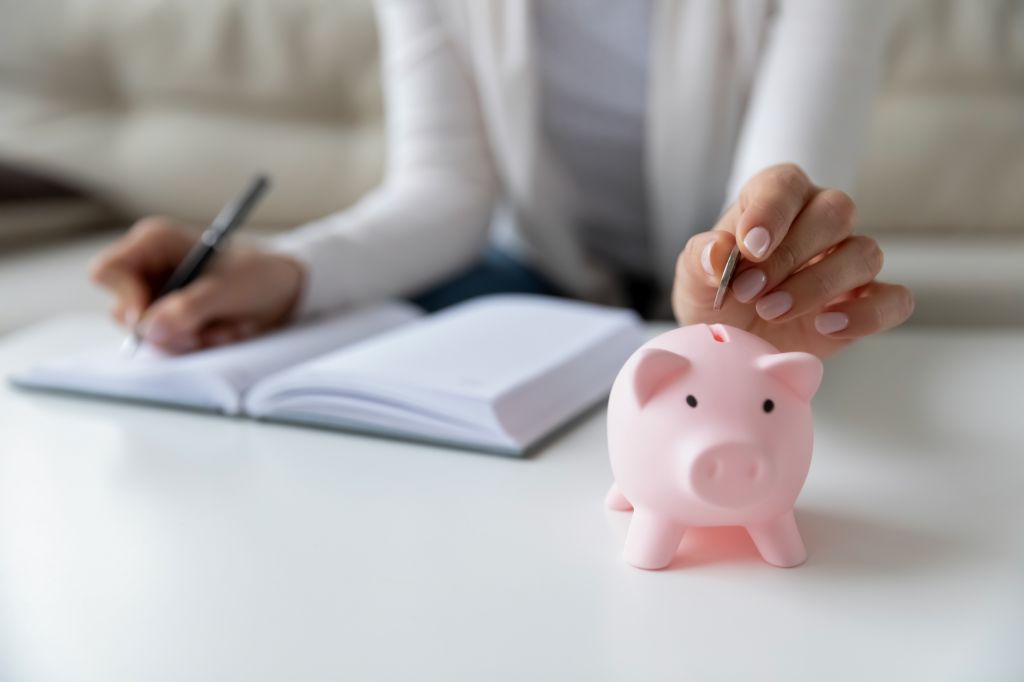 Thrifty woman sit at table hold pen writing daily expenses in diary put coin in pink piggy bank close up.