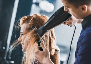 Young hairdresser drying customers hair with round brush at hair salon.