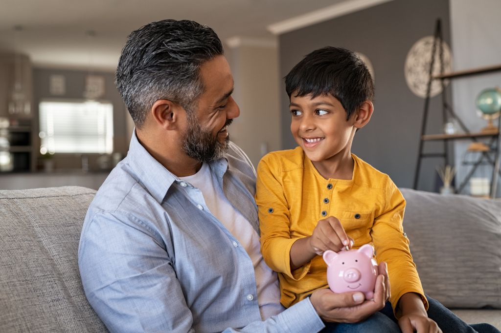 Indian father and smiling son putting coin into piggy bank