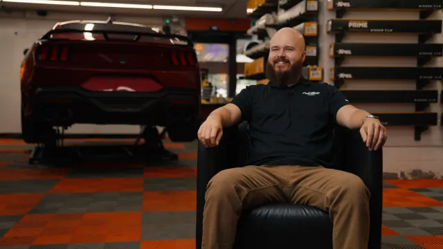 FullMoon Autoworks founder, Erik Wigger, sits smiling in his specialty automotive shop, with a car on a lift and window-tinting boxes visible in the background.