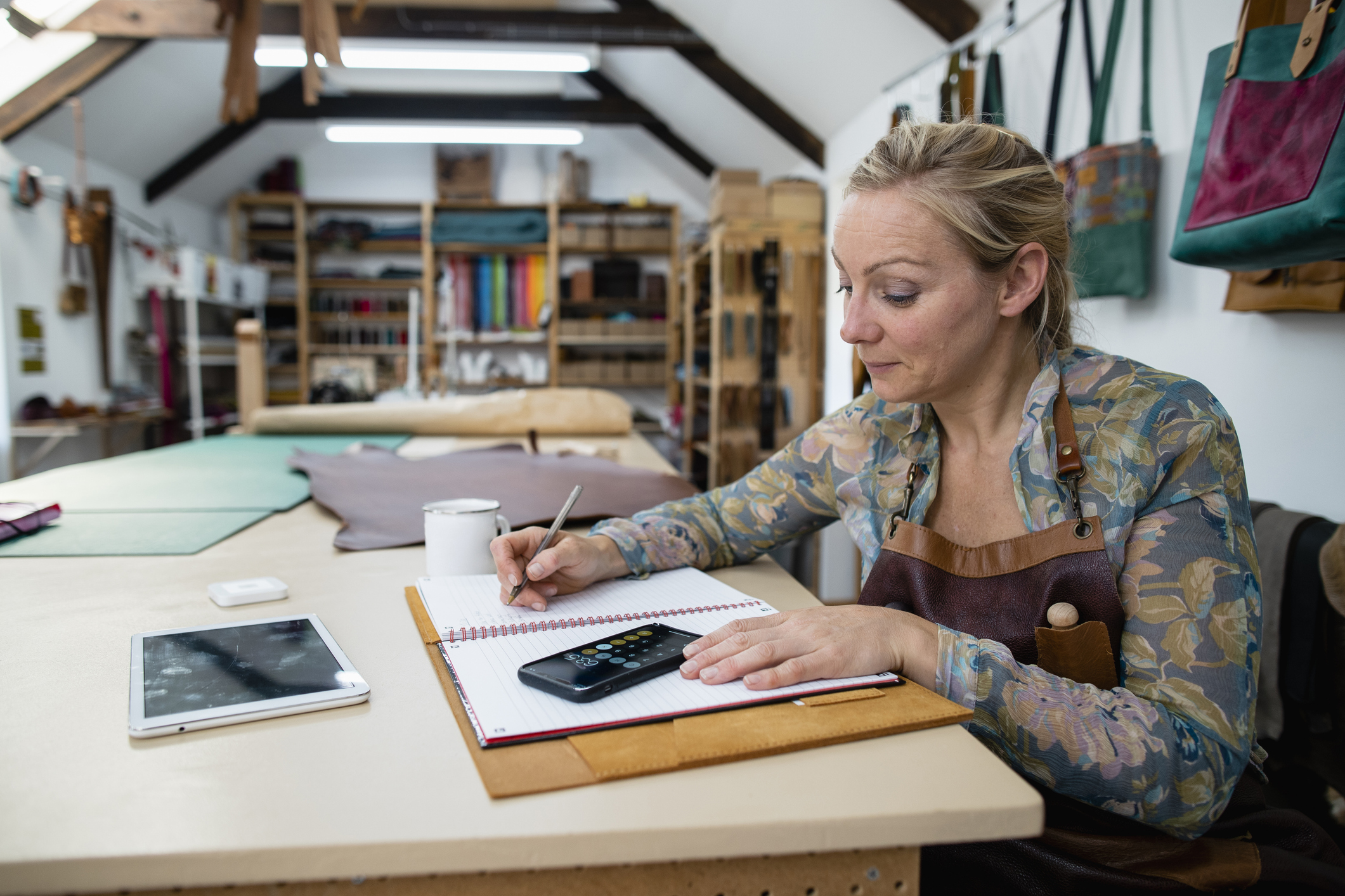 A tanner sitting in her tannery studio at a workbench, updating her account books with profits from sales she has made.