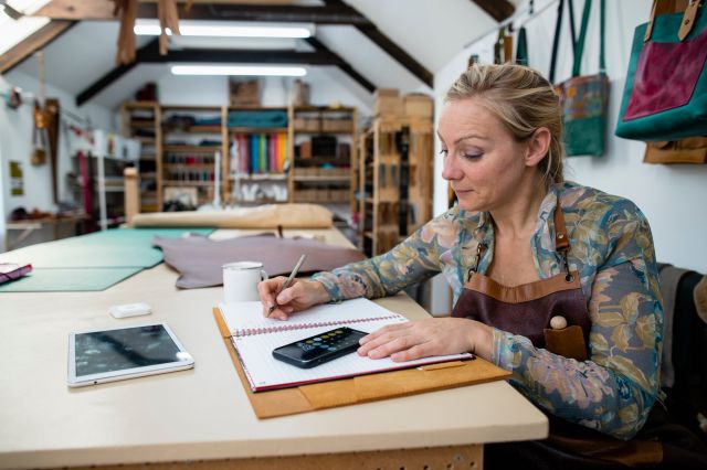 A tanner sitting in her tannery studio at a workbench, updating her account books with profits from sales she has made.