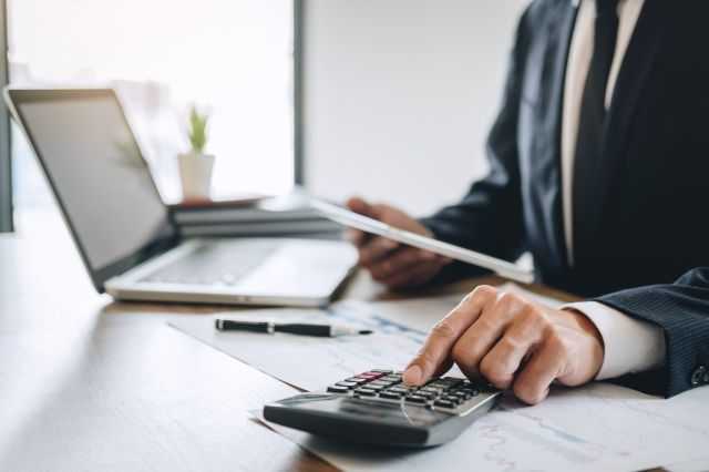 Businessman working at a desk with papers, a laptop open, and he is typing on a calculator.