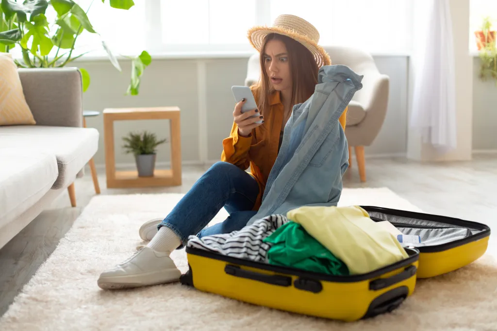 Female with hat packing her suitcase for trip while looking at phone