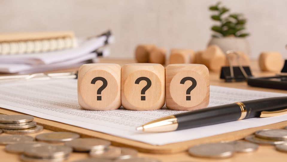 Three wooden cubes with question marks are placed on financial documents, surrounded by coins, a pen, and office supplies, representing financial uncertainty, decision-making, and economic challenges.