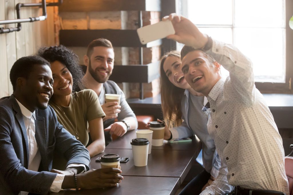 A group of five young adults sitting at a café table, smiling and taking a selfie together. They are casually dressed and appear to be enjoying coffee and each other’s company. The setting is bright with natural light coming through large windows.