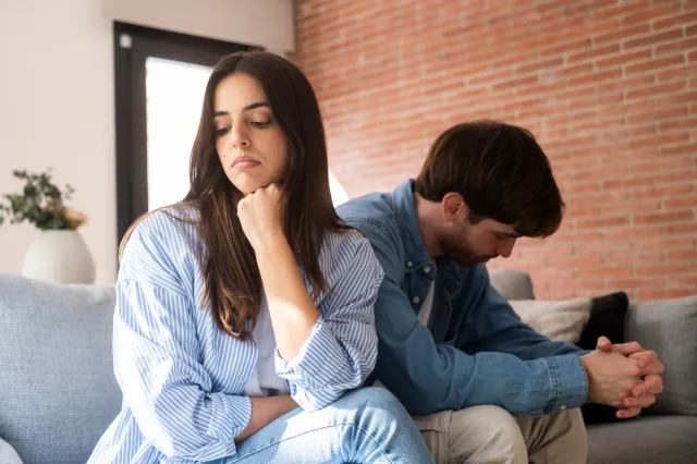 A couple sitting on a grey couch facing away from each other, appearing upset.