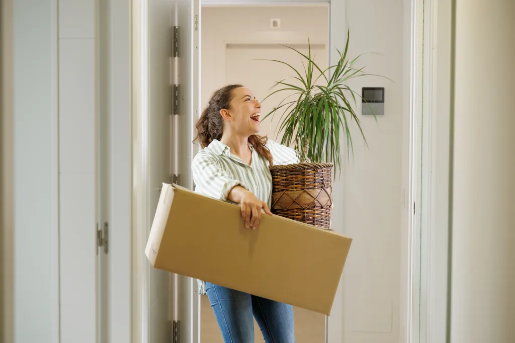 Smiling young woman carrying a moving box and plant as she moves into her first home.