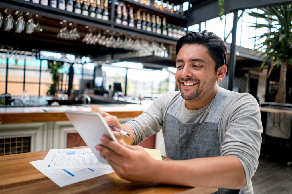 A smiling aproned business owner holds his tablet while reviewing payroll documents at a bar counter.