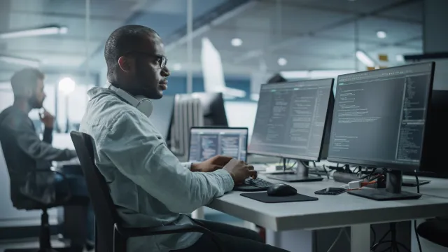 A business leader sits in an office at his desk looking at monitor screens
