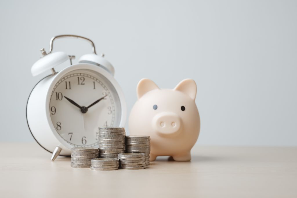 An illustrative image of clock and piggy bank sitting on a desk.