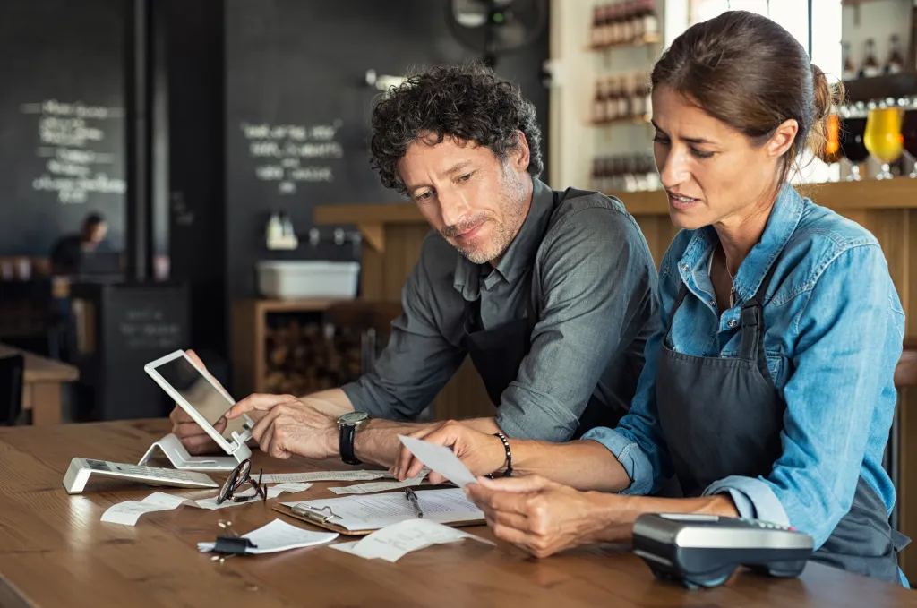 Man and woman sitting in cafeteria discussing finance for the month. Stressed couple looking at bills sitting in restaurant wearing uniform apron.