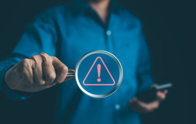 Man holding a phone in one hand and a magnifying glass with a caution symbol in the other.