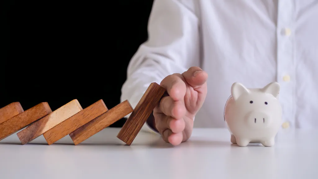 Hand stopping falling dominos next to piggy bank, symbolizing financial protection and savings stability