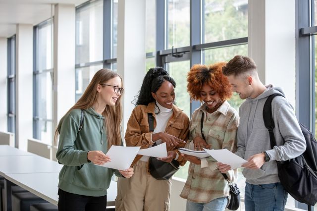 Four smiling university students reviewing documents together in a bright campus building, each carrying a backpack.