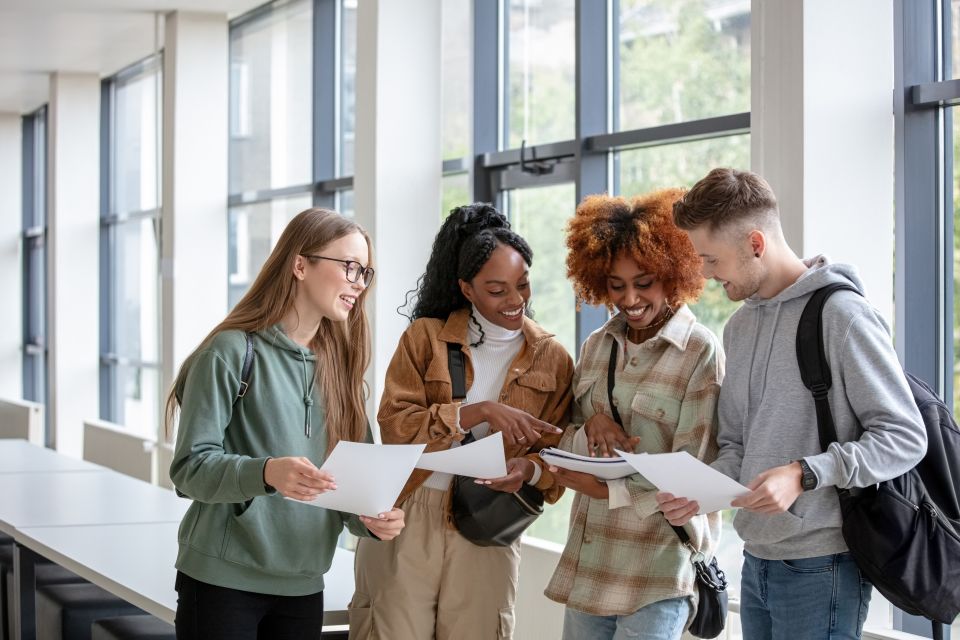 Four smiling university students reviewing documents together in a bright campus building, each carrying a backpack.