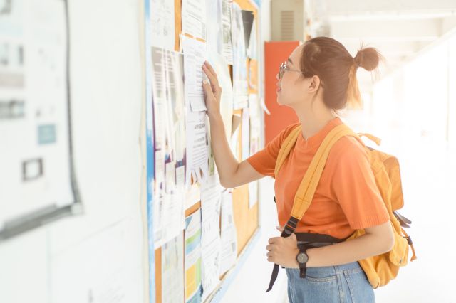 A young woman wearing glasses, an orange top, and a yellow backpack reads job postings on a bulletin board in a bright hallway.