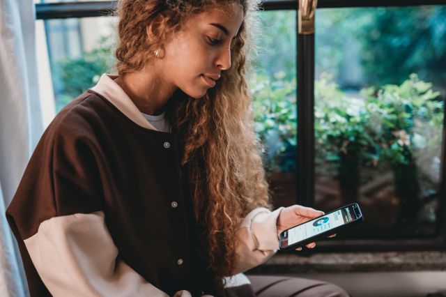 A young person with long curly hair sits by a window, looking down at a smartphone displaying a financial app, with lush greenery visible in the background.